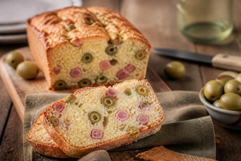 Cake jambon olive moelleux tranché sur une planche en bois, avec des olives vertes et du gruyère râpé visibles