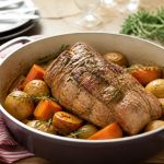 Rôti de veau en cocotte en fonte dorée, entouré de légumes et d'herbes fraîches, sur une table en bois rustique