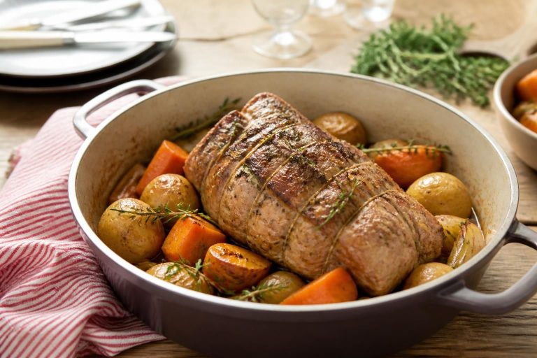 Rôti de veau en cocotte en fonte dorée, entouré de légumes et d'herbes fraîches, sur une table en bois rustique