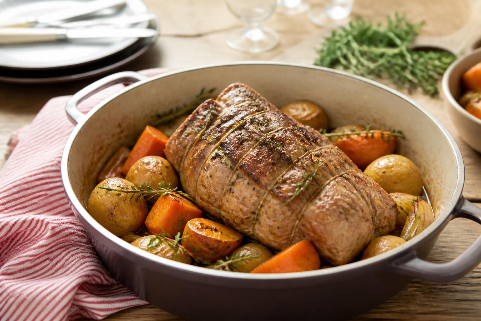 Rôti de veau en cocotte en fonte dorée, entouré de légumes et d'herbes fraîches, sur une table en bois rustique